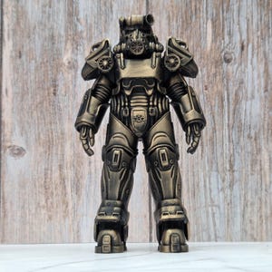 May include: A bronze-colored figurine of a power armor suit, featuring intricate details and a weathered finish. The suit has a helmet with a respirator, shoulder pads, and segmented armor plating. The figure stands upright, showcasing the full design.