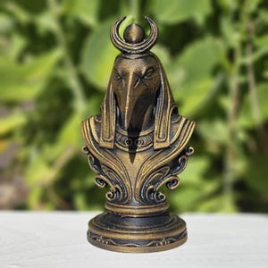 May include: A bronze-colored bust of an Egyptian deity with a bird-like head, adorned with a crescent moon and sun disc. The sculpture features intricate details and a decorative base, showcasing a blend of ancient artistry and craftsmanship.