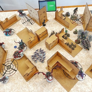 以下が含まれることがあります： ミニチュアフィギュアと地形を備えた卓上ウォーゲームのセットアップ。茶色の段ボール構造、円形プラットフォーム、さまざまな塗装されたフィギュアが含まれています。デジタルタイマーが時間を表示します。ゲームは進行中のようです。