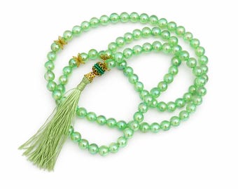 May include: A green beaded necklace with a tassel. The necklace features iridescent green beads, gold-colored accents, and a green tassel. The beads are strung together, forming multiple strands. The tassel is made of fine, light green threads.