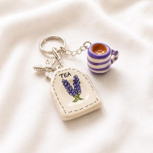 Könnte beinhalten: Ein Metallschlüsselanhänger mit einem silbernen Bienenanhänger, einer kleinen Keramiktasse mit violett-weißen Querstreifen und einem cremefarbenen Keramik-Teebeutelanhänger mit violetten Lavendelzweigen und dem schwarzen Text TEA.