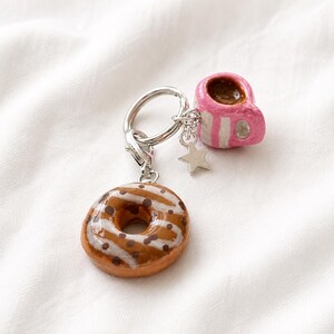 Könnte beinhalten: Ein Schlüsselanhänger aus silberfarbenem Metall mit einem braun glasierten Donut-Anhänger mit weißen Zuckergussstreifen und braunen Streuseln, ergänzt durch einen kleinen rosa-weiß gestreiften Tassenanhänger und einen silbernen Stern.