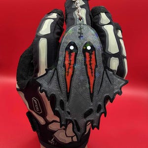 May include: A gray ghost-shaped keychain with red eyes and a bat-like wing design. The keychain is attached to a silver keyring. The background is a solid red color.