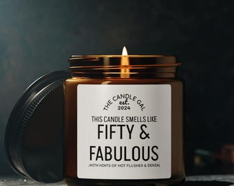 Puede incluir: Una vela encendida en un tarro de cristal marrón con tapa dorada. La etiqueta dice "THIS CANDLE SMELLS LIKE FIFTY & FABULOUS" con el texto "THE CANDLE GAL est. 2024". La tapa está a un lado.
