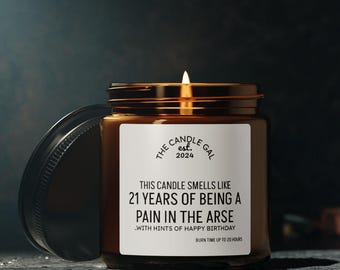 Puede incluir: Una vela de vidrio marrón encendida con tapa dorada y etiqueta blanca. La etiqueta dice "THIS CANDLE SMELLS LIKE 21 YEARS OF BEING A PAIN IN THE ARSE...WITH HINTS OF HAPPY BIRTHDAY." La vela es de "The Candle Gal est. 2024."