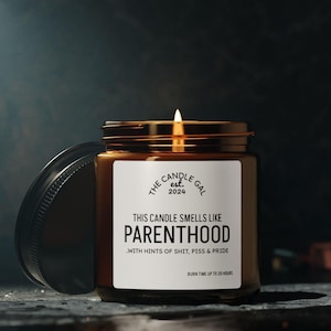 May include: A lit brown glass candle jar with a gold lid and a white label that reads "THIS CANDLE SMELLS LIKE PARENTHOOD" with the text "WITH HINTS OF SHIT, PISS & PRIDE". The candle is from "THE CANDLE GAL est. 2024".