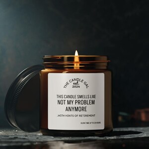 Może przedstawiać: Zapalona świeca w bursztynowym szklanym słoiku z czarną pokrywką. Biała etykieta zawiera napis: THE CANDLE GAL est. 2024, THIS CANDLE SMELLS LIKE NOT MY PROBLEM ANYMORE, WITH HINTS OF RETIREMENT, BURN TIME UP TO 20 HOURS. Płomień świecy jest widoczny.