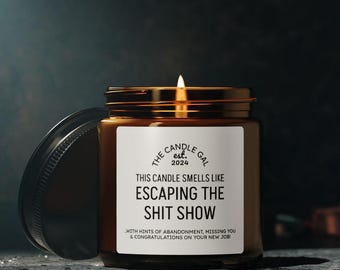 May include: A lit brown glass candle jar with a gold lid and a white label. The label reads "The Candle Gal est. 2024" and "This candle smells like escaping the shit show." The candle is on a dark surface.