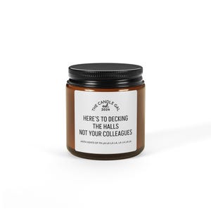 May include: A brown glass jar candle with a black lid. The label reads "THE CANDLE GAL 2024" and "HERE'S TO DECKING THE HALLS NOT YOUR COLLEAGUES" with "WITH HINTS OF FA LA LA LA LA, LA LA LA LA" below.