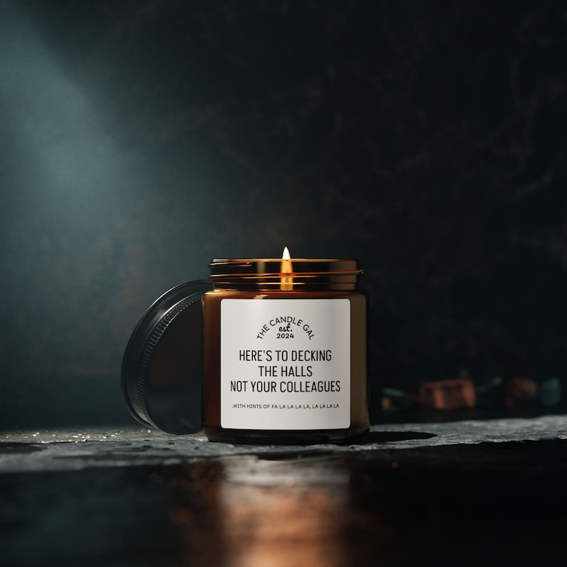 May include: A lit candle in a brown glass jar with a gold lid. The label reads "Here's to Decking the Halls Not Your Colleagues." The candle is from "The Candle Gal est. 2024" with the text "With hints of pa la la la la la la la la."