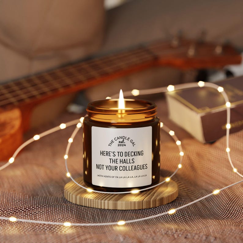 May include: A lit candle in a brown glass jar with a gold lid, sitting on a wooden coaster. The label reads "The Candle Gal est. 2024" and "Here's to decking the halls not your colleagues." Fairy lights and a ukulele are in the background.