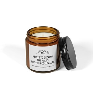 May include: A brown glass jar candle with a black lid. The candle has a white label with the text "HERE'S TO DECKING THE HALLS NOT YOUR COLLEAGUES" and "WITH HINTS OF FA LA LA LA LA LA LA LA".
