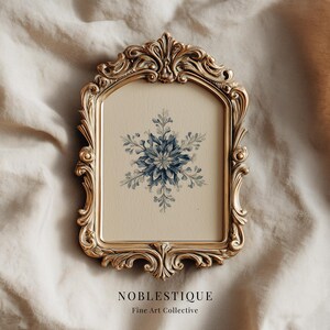 Puede incluir: Marco dorado ornamentado con una impresión de acuarela de un copo de nieve azul sobre un fondo crema. El marco tiene intrincados adornos y una estética vintage. El texto "NOBLESTIQUE Fine Art Collective" está impreso en la parte inferior.