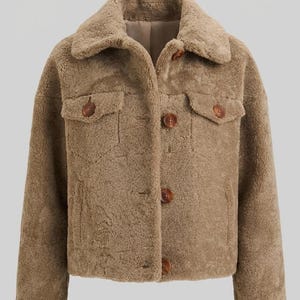 Puede incluir: Una chaqueta de piel sintética color canela con cuello, mangas largas y cierre frontal de botones. La prenda cuenta con dos bolsillos en el pecho con solapas abotonadas y botones circulares de color marrón oscuro, con textura afelpada.