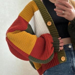 May include: A colourful, handmade crochet cardigan with a patchwork design. The sweater features blocks of mustard yellow, red, white, and olive green. It has large wooden buttons and a relaxed fit. The sleeves are a balloon style.