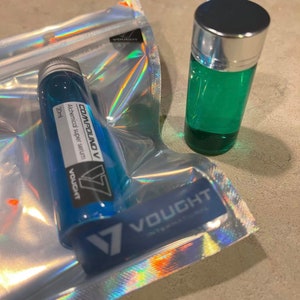 以下が含まれることがあります： 銀色のキャップと白いラベルに「COMPOUND V Alchemical super serum 20ml VOUGHT」と書かれた青いガラス瓶。瓶は、ホログラフィックな虹模様の透明なビニール袋に入っています。背景には、銀色のキャップが付いた小さな緑色のガラス瓶があります。