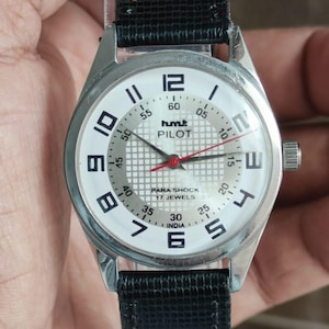 May include: A silver wristwatch with a white face, black numerals, and hands. The watch face displays "hmt PILOT" and "PARA SHOCK 17 JEWELS INDIA". The watch has a black leather strap.