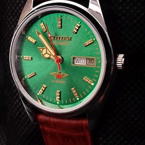 Pode incluir: Um relógio de pulso automático Citizen com mostrador verde e detalhes dourados. O relógio tem um ponteiro de segundos vermelho e um visor de data mostrando "QUA 30". Possui uma pulseira de couro castanho.