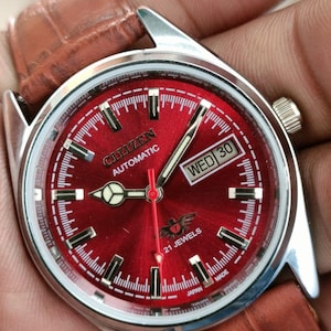 May include: A Citizen automatic wristwatch with a red dial and a brown leather strap. The watch face displays the day and date, with the text "WED 30". The watch has silver hands and hour markers. The watch is marked "21 JEWELS" and "JAPAN MADE".