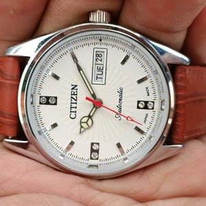 Op de afbeelding: Citizen automatisch polshorloge met een witte wijzerplaat, zilveren kast en bruine leren band. De wijzerplaat toont de dag en datum, met een rode secondewijzer. Het horloge is gemarkeerd met "Made in Japan."