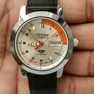 Puede incluir: Reloj automático Citizen plateado con correa de cuero negro. La esfera del reloj es plateada con detalles en naranja y negro, y muestra el día de la semana. Caja plateada, correa de cuero negro.