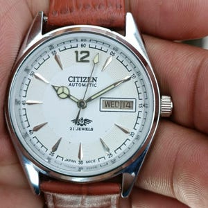 Può includere: Un orologio da polso automatico Citizen con quadrante bianco, lancette argentate e cinturino in pelle marrone. Il quadrante mostra il giorno e la data, con la scritta "CITIZEN AUTOMATIC" e "21 JEWELS". L'orologio è tenuto in una mano.