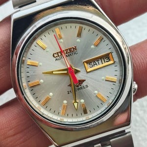 Op de afbeelding: Zilverkleurig Citizen automatisch horloge met een lichtgrijze wijzerplaat. Het horloge heeft goudkleurige uurmarkeringen en wijzers, met een rode secondewijzer. De dag en datum worden weergegeven in een klein venster. De horlogeband is ook zilverkleurig.