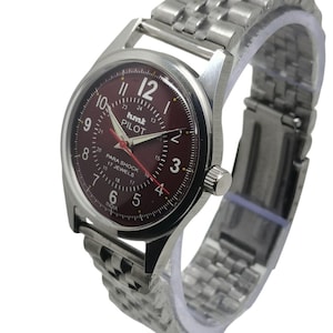 May include: A silver wristwatch with a burgundy dial and a stainless steel strap. The watch face displays the text "hmt PILOT" and "PARA SHOCK 17 JEWELS". The watch hands are red. The watch has a classic design.