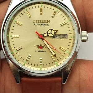 Puede incluir: Un reloj de pulsera Citizen Automatic con esfera dorada, caja plateada y correa de cuero marrón. La esfera muestra el día y la fecha, con la marca y "21 Jewels" visibles. El reloj tiene un diseño clásico.