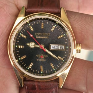 May include: A gold-toned Citizen automatic wristwatch with a black face and brown leather band. The watch face displays the day and date, with gold markers and hands. The watch has a red second hand and the text "Citizen Automatic" is visible.