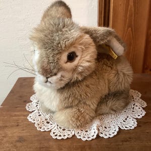 May include: A plush brown and white rabbit toy with a white lace doily underneath. The rabbit has a long, fluffy tail and a small, black nose.