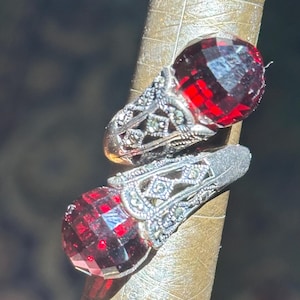 May include: Two silver rings with large, faceted, deep red gemstones. The rings are on a ring sizer. The silver bands have an ornate, filigree design with small, sparkling accents. The gemstones are a vibrant, eye-catching red.