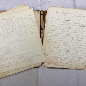May include: An open antique cookbook with yellowed, aged paper pages featuring handwritten cursive recipes in black ink. The left page lists ingredients for Baked Oysters and Berry Pudding, while the right page details a Potato Scallop recipe.