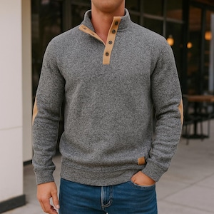 Könnte beinhalten: Ein grauer Pullover mit hohem Kragen und beigen Akzenten an Kragen, Ellbogenpatches und Knöpfen. Der Pullover hat eine strukturierte Optik und wird mit einer blauen Jeans kombiniert. Ein kleines braunes Etikett ist unten rechts sichtbar.