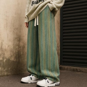 May include: Wide-leg, vertically striped pants in shades of green and tan. The pants have a drawstring waist and are paired with white sneakers with black accents. The person is wearing a light-colored sweater with the word "Bears" on it.