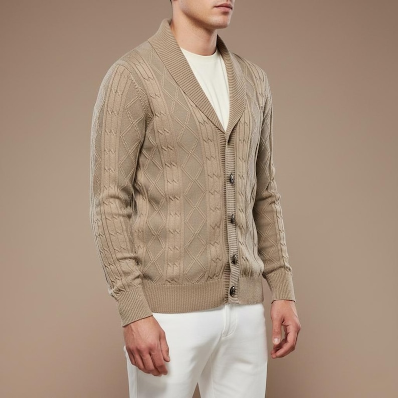 K&ouml;nnte beinhalten: Beigefarbene Strickjacke mit Rautenmuster und Schalkragen. Der Pullover mit Kn&ouml;pfen hat lange &Auml;rmel. Die Person auf dem Bild tr&auml;gt ein wei&szlig;es T-Shirt und eine wei&szlig;e Hose.