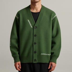Könnte beinhalten: Grüner Cardigan mit V-Ausschnitt und Knopfverschluss. Weiße Nahtdetails sind an den Ärmeln und der Brust zu sehen. Die Knöpfe sind schwarz. Das Model trägt ein schwarzes T-Shirt darunter.