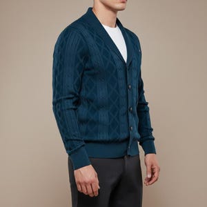 K&ouml;nnte beinhalten: T&uuml;rkisfarbene Strickjacke mit Zopfmuster, V-Ausschnitt und Knopfverschluss. Der Pullover hat ein Rautenmuster und wird &uuml;ber einem wei&szlig;en T-Shirt getragen. Die &Auml;rmel sind lang und die Strickjacke ist aus weichem, strukturiertem Material.