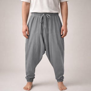 May include: Gray harem pants with a drawstring waist and elastic cuffs. The pants have a relaxed fit and a drop-crotch design. The fabric appears to be a lightweight material, suitable for casual wear. The pants are shown on a person.