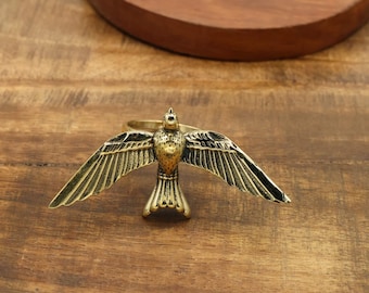 May include: A brass bird ring with outstretched wings, set on a wooden surface. The ring has detailed feather etching and a circular band. The bird is the focal point, with a vintage aesthetic. The background includes a wooden plate.