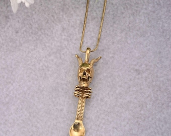May include: Gold-colored pendant necklace featuring a skull with horns and a spoon-shaped design. The pendant is suspended from a delicate chain. The skull has detailed features, and the spoon has a textured handle.
