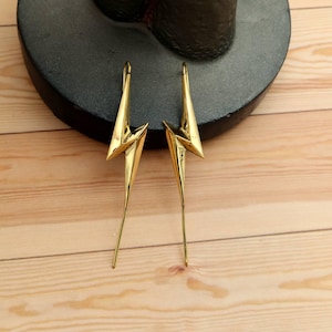 May include: A pair of gold-colored earrings in a lightning bolt design. The earrings have a pointed top and a long, slender bottom. They are displayed on a dark gray surface against a light brown wooden background.