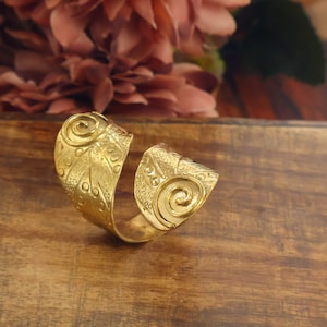 Può includere: Un anello aperto color oro caratterizzato da una fascia larga con superficie testurizzata. Il design presenta due motivi a spirale prominenti alle estremità, completati da un motivo a piccoli punti in rilievo e una finitura granulata.