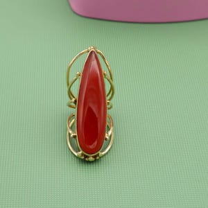 May include: A gold-colored ring featuring a large, teardrop-shaped, red gemstone. The ring has an ornate, openwork design around the stone and band. The ring is set against a green background.