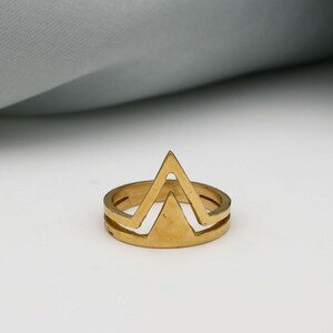 May include: A gold-coloured ring with a geometric mountain-like design. The ring features a double band and a triangular peak, creating a modern and minimalist aesthetic. The ring is likely made of metal.
