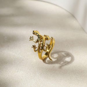 May include: A gold-colored ring in the shape of a lizard. The lizard has detailed features, including tiny feet and a textured body. The ring is designed to wrap around the finger, with the lizard's tail curving around the band.