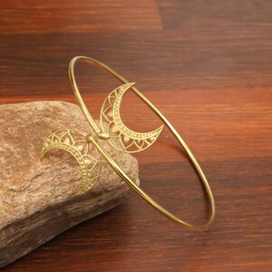 May include: A gold-toned metal cuff bracelet featuring two crescent moon motifs at the open ends. Each crescent moon is decorated with an intricate, filigree-style cutout pattern. The bracelet has a thin, circular band design.