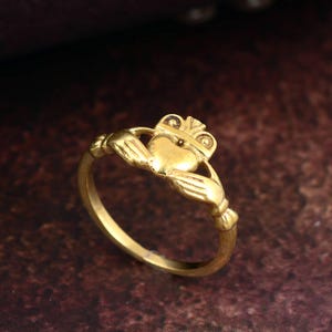 May include: A gold Claddagh ring, with a heart held by two hands and a crown. The ring is set against a dark, textured background, highlighting the intricate details. The ring's band is smooth and rounded. The ring is a symbol of love, loyalty, and friendship.