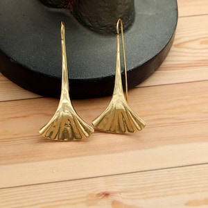 May include: A pair of gold-coloured earrings with a fan-shaped design. The earrings have a long, slender hook and a textured, petal-like bottom. The earrings are displayed on a wooden surface.