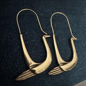May include: A pair of gold-colored swan-shaped earrings. The earrings feature a curved design with a swan's head and detailed wing feathers. The earrings are displayed against a dark blue background.